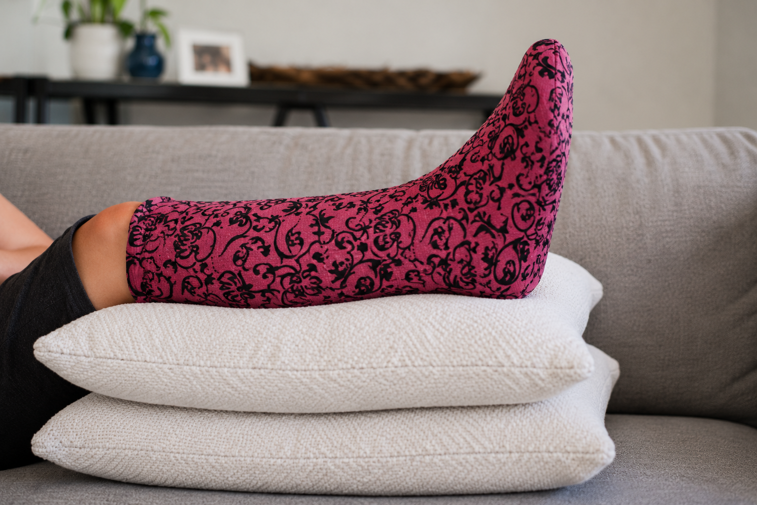 Leg cast elevated on stacked pillows while resting on a couch, covered with a pink and black paisley CastCoverz! cast cover to help protect the cast and provide comfort during orthopedic recovery.
